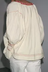 Camisa color crudo de cuello redondo con escote pico y lazada. Manga larga acabada en volantes. Detalle de bordados combinados a contraste y puntilla a tono.