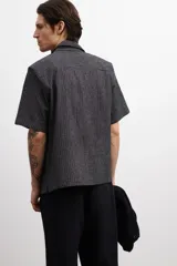 Camisa de manga corta color gris oscuro con rayas verticales, cuello resort y bolsillo de parche en el pecho. Corte recto y cierre con botones.