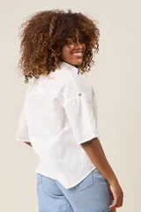 Camisa color beige claro, de corte clásico, con cuello, botones frontales, bolsillo en el pecho y mangas tres cuartos con presilla para sujetar.