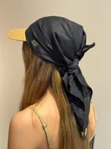 Pañuelo de lino negro anudado en la nuca, usado sobre una gorra de visera color camel. El pañuelo tiene un bordado de iniciales en hilo claro.
