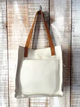 Bolso tote de cuero color blanco hueso, con asas cortas de cuero marrón y un bolsillo exterior grande en el frente.