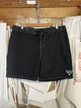 Short de baño negro de largo medio (18 pulgadas) con costuras reforzadas en hilo blanco contrastante. Presenta cordón de ajuste en la cintura y un pequeño logo estampado en la pierna derecha.