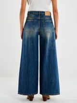 Jeans de mezclilla azul oscuro con lavado vintage y efecto degradado en las piernas. Diseño de tiro medio con pierna muy ancha (palazzo o culotte ancho) y costura central vertical en el frente de cada pierna.