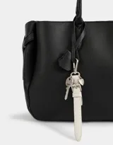 Cartera shopper de cuerina color negro con manijas superiores y charm a contratono. Tiene broche con botón para cerrar y cinturones laterales.