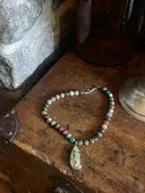 Collar de piedras naturales redondas, con tonos verdes, marrones, rojizos y rosados, engarzadas en una cadena plateada. Incluye un dije colgante en forma de gota engarzado en metal plateado, con una piedra iridiscente.