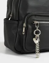 Mochila urbana negra de cuerina con bolsillo frontal con cierre y charm colgante de cadena con dije de corazón y candado. Tiene correas regulables, manija superior, cierre superior, doble bolsillo frontal (uno con charm metálico) y bolsillos laterales.