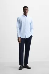 Camisa celeste a rayas verticales blancas, de corte recto, con cuello italiano abierto, manga larga con puños abotonados y cierre frontal con botones.