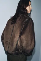 Cazadora de estilo aviador con cuello subido, manga larga y cierre frontal asimétrico con cremallera. Presenta un diseño de doble faz con exterior de efecto ante y forro interior de borreguillo a tono, con detalles de ribetes en cuero sintético.