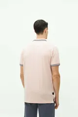 Chomba de piqué color rosa claro, de corte clásico, con cuello y mangas con ribete a rayas en blanco, gris y negro. Tiene tapeta de dos botones y logo bordado en el pecho.