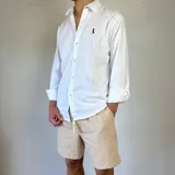 Camisa blanca de algodón con cuello clásico, manga larga y cierre frontal con botones. Tiene un pequeño logo bordado en el pecho.