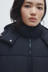 Campera puffer larga negra con capucha, cuello elevado, cierre frontal, bolsillos insertados y elástico oculto en los puños.