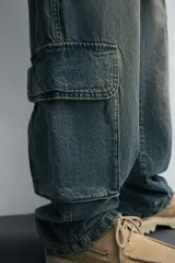 Pantalón de denim color beige, corte relaxed fit, con bolsillos laterales tipo cargo con solapa y cierre de botón. Cierre frontal con botón y cremallera.
