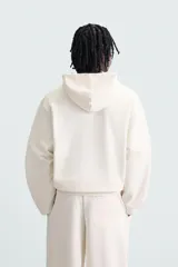 Sudadera con capucha color crudo, de corte relajado, confeccionada en tejido de algodón con interior rizado. Cuenta con cuello con capucha, manga larga, bolsillo frontal tipo canguro y acabados en rib.