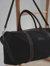 Bolso de lona color negro con detalles en cuero marrón, doble asa de mano y correa de hombro ajustable y extraíble.