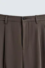 Pantalón negro de corte ancho con cintura elástica, pliegues frontales, bolsillos laterales y traseros de vivo. Tiene detalle de hebilla metálica en la trabilla y cierre frontal con cremallera y botón.