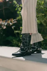 Bota de caña media color negro, estilo texana, con punta fina, tachas plateadas y apliques de hebillas metálicas con cadena.