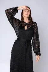 Vestido largo negro de encaje calado, con mangas largas abullonadas y cuello redondo con abertura en forma de lágrima. Incluye un lazo de tela para ajustar a la cintura.