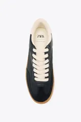 Zapatillas deportivas de mujer con combinación de materiales en negro, blanco y marrón. Cierre con cordones y suela en contraste color caramelo.