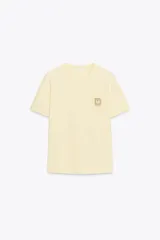 Camiseta marrón de algodón de manga corta y cuello redondo. Presenta un bolsillo en el pecho con un broche metálico dorado con diseño intrincado.