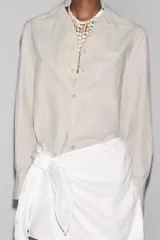 Camisa de manga larga color crudo, confeccionada en hilatura de algodón. Tiene cuello solapa, bolsillo de plastrón en el pecho, bajo redondeado y cierre frontal con botones.