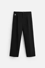 Pantalón negro de corte ancho con cintura elástica, pliegues frontales y detalle de hebilla metálica en la trabilla. Tiene bolsillos delanteros y traseros de vivo, y cierre frontal con cremallera y botón.