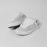 Zapatillas bajas de lona color gris claro, con cordones blancos gruesos y suela de goma blanca. Presentan el logo de la marca en el lateral exterior.