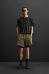 Shorts deportivos color verde oliva con calza interior negra, de tejido técnico ligero y elástico. Cuenta con cintura elástica ajustable con cordón, bolsillos laterales con cierre de cremallera, bajo con aberturas laterales y detalle de logo estampado.