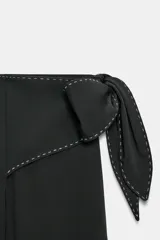 Pantalón negro de tiro medio con cremallera lateral oculta, corte ancho y lazo decorativo en la cadera. Detalle de pespuntes blancos a contraste.
