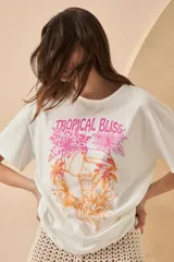 Remera blanca de algodón con estampado de un tucán, palmeras y la frase 'Tropical Bliss' en tonos naranja y rosa.