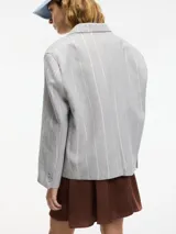 Blazer gris a rayas verticales blancas con lurex, con cuello de solapa, cierre con botón único y tajo en la espalda. Tiene un bolsillo de ojal en el pecho y bolsillos con solapa a la altura de la cadera.