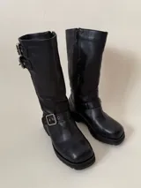 Bota de caña media de cuero vacuno color marrón oscuro, con punta cuadrada, cierre lateral y suela track negra. Presenta detalles de hebillas metálicas plateadas en la caña y en la parte inferior del empeine.