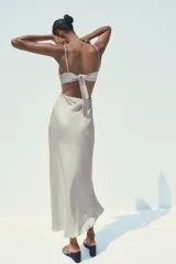Vestido midi de satén color gris claro, con escote recto, tirantes finos regulables, cuerpo tipo pañuelo con terminación en pico, espalda descubierta con lazo y cierre lateral con cremallera oculta.