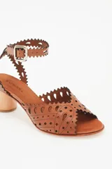 Sandalia de cuero color caramelo con taco bajo de madera. Presenta un diseño calado con bordes festoneados y perforaciones, y tira ajustable con hebilla en el tobillo.