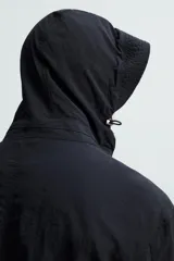 Parka negra de tejido técnico ligero, corte regular, cuello alto con capucha ajustable, manga larga con puños ajustables, cintura ajustable con cordón interno, bolsillos de solapa en la cadera, abertura en la espalda y cierre frontal con cremallera oculta y botones a presión.