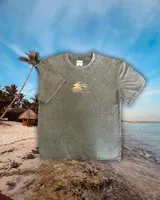 Remera de manga corta, color verde musgo con efecto gastado (acid wash). Estampado en la espalda con un diseño de atardecer tropical con palmeras y texto circular que dice "SUNSET", "TROPICAL" y "PALM BEACH".