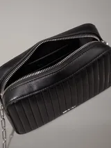 Cartera bandolera Calvin Klein negra con diseño acolchado de líneas verticales, correa de cadena plateada, cierre superior con cremallera y logo metálico frontal.
