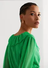 Vestido corto verde con textura, diseño evasé, cuello escotado con volantes, manga larga con puños elásticos, fruncido elástico en la cintura, volante en el bajo y cierre de lazo en el cuello.