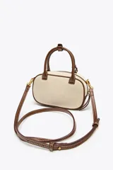Bolso mini color crudo con detalles en marrón. Tiene asa de mano y doble asa bandolera extraíble, una en tejido tipo canvas y otra con pespunte. Incluye charm con piezas metálicas y bolsillo interior con cremallera.