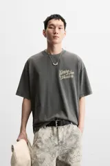 Camiseta gris oscuro con efecto lavado, cuello redondo y manga corta. Tiene un bolsillo en el pecho y estampados en el frente y la espalda.