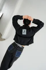 Campera de jean negra tipo bomber con cuello redondo y cierre frontal con botones metálicos. Presenta un parche rectangular con diseño gráfico en la espalda.