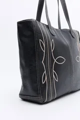 Cartera tipo tote bag en símil cuero color marrón oscuro, con bordados decorativos en blanco que evocan un estilo western. Cuenta con asas largas para llevar al hombro.