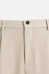 Pantalón gris de corte ancho con cintura elástica, pliegues frontales, bolsillos laterales y traseros de vivo. Incluye detalle de hebilla metálica en la trabilla y cierre frontal con cremallera y botón.