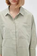 Camisa color verde claro, de corte holgado, con cuello clásico, cierre frontal con botones, mangas 3/4 y dos bolsillos de parche en el pecho.