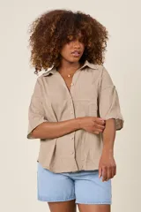 Camisa color beige claro, de corte clásico, con cuello, botones frontales, bolsillo en el pecho y mangas tres cuartos con presilla para sujetar.