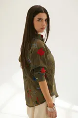 Camisa beige de manga larga con cuello clásico y cierre frontal con botones. Presenta bordados con motivos florales y geométricos en tonos azules, rojos y verdes.