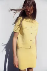 Chaqueta de punto color amarillo pálido, con cuello redondo y manga corta. Cierre frontal con botones dorados y bolsillos de parche en el pecho.