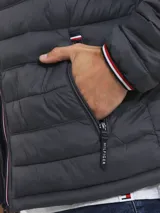Chaqueta acolchada gris de poliamida con cuello alto, cierre de cremallera, dos bolsillos delanteros con cremallera y logo de Tommy Hilfiger bordado en el pecho.