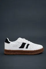 Zapatilla deportiva blanca de caña baja, con detalles en negro en el talón y dos franjas negras inclinadas en el lateral. Posee una suela gruesa de goma color marrón.