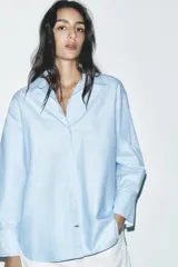 Camisa celeste de corte clásico, con cuello solapa, manga larga, bajo asimétrico y cierre frontal con botones.