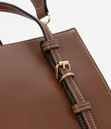 Bolso satchel mediano color marrón, confeccionado en poliuretano, con correa de mano con detalles de hebilla y costura, asa larga desmontable con gorgorão y cierre por cremallera.
